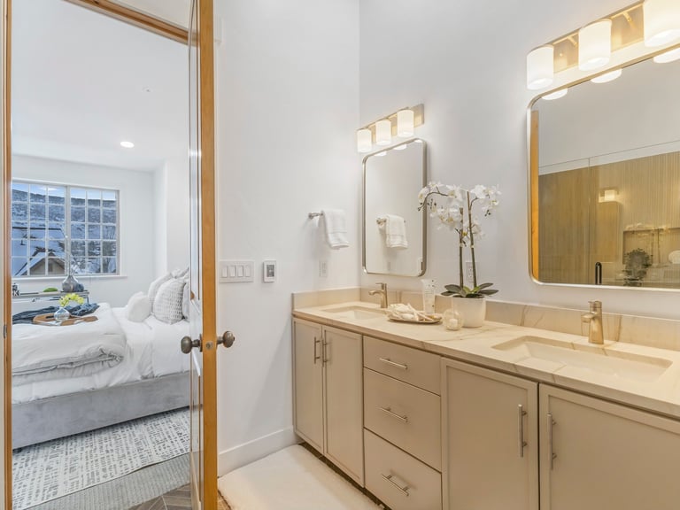 Modern luxury bathroom with dual vanity, brass-framed mirrors, and view of bedroom with city windows