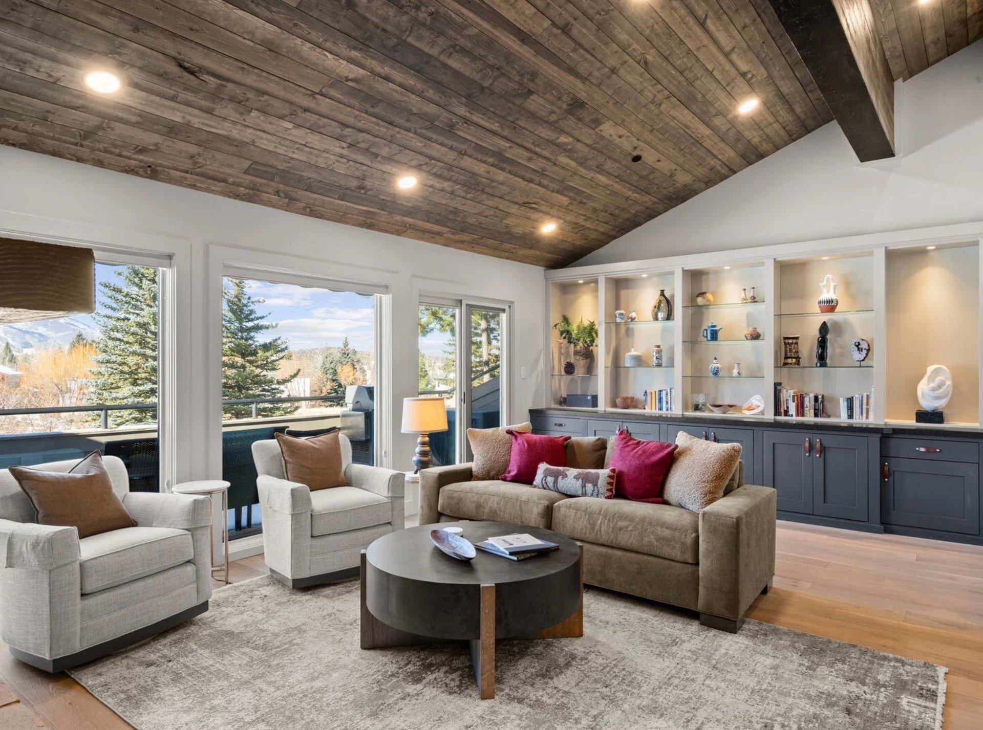 Modern luxury living room with wood ceiling, large windows, beige sofa, armchairs, round black coffee table, and built-in shelving