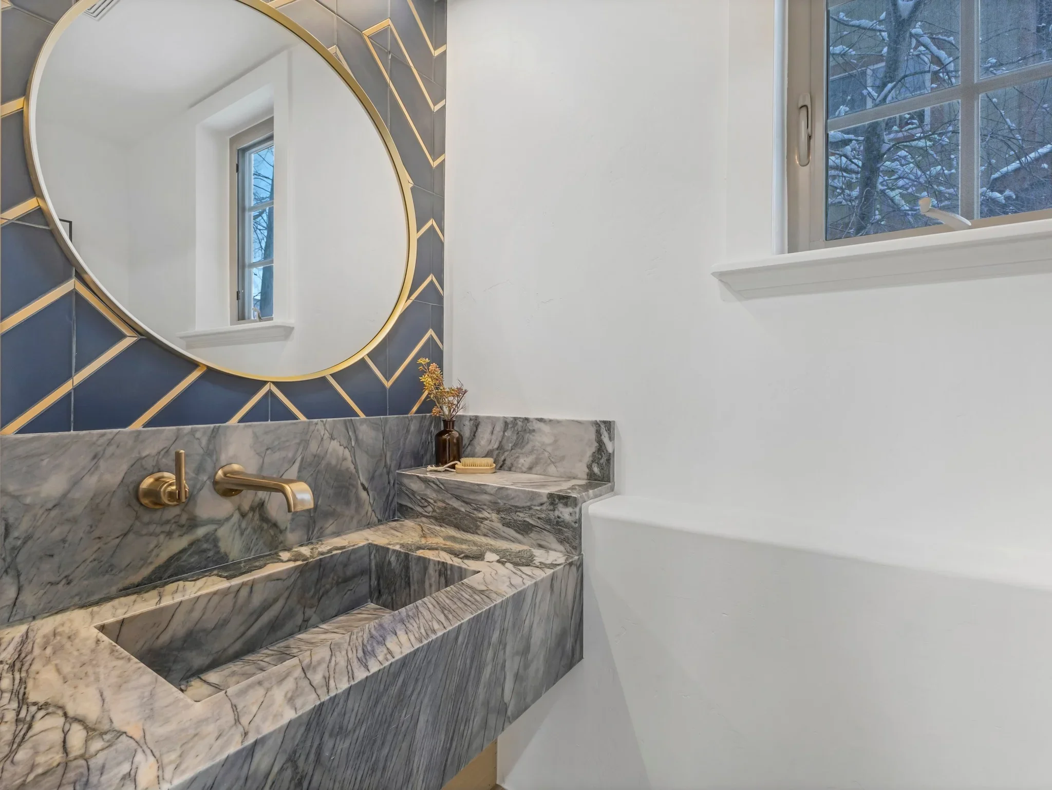 Marble vanity with dual sinks, brass fixtures, circular mirror, and geometric blue accent wall in modern bathroom