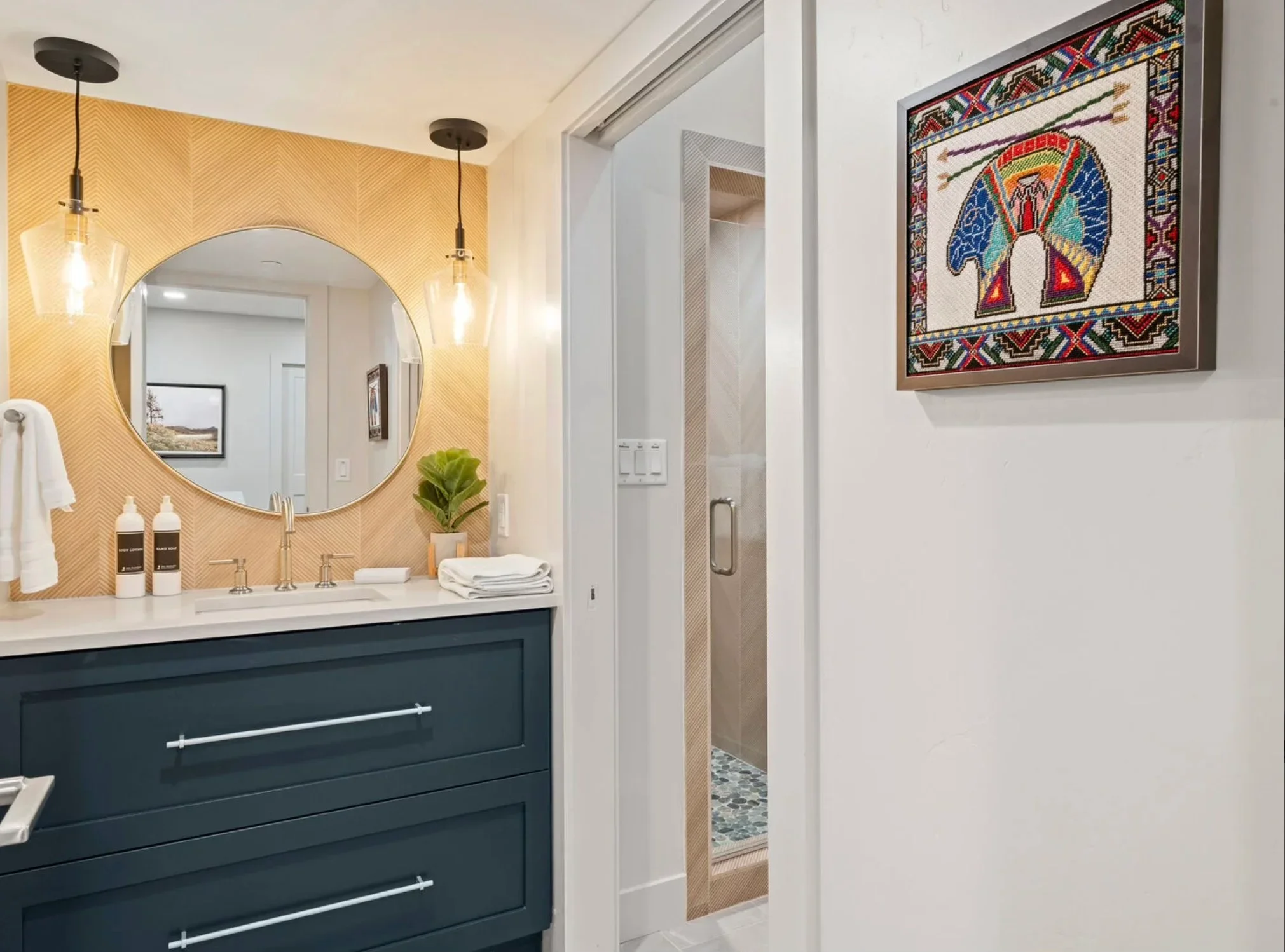 Bathroom with dark teal vanity, gold herringbone tile, round mirror, and pendant lights