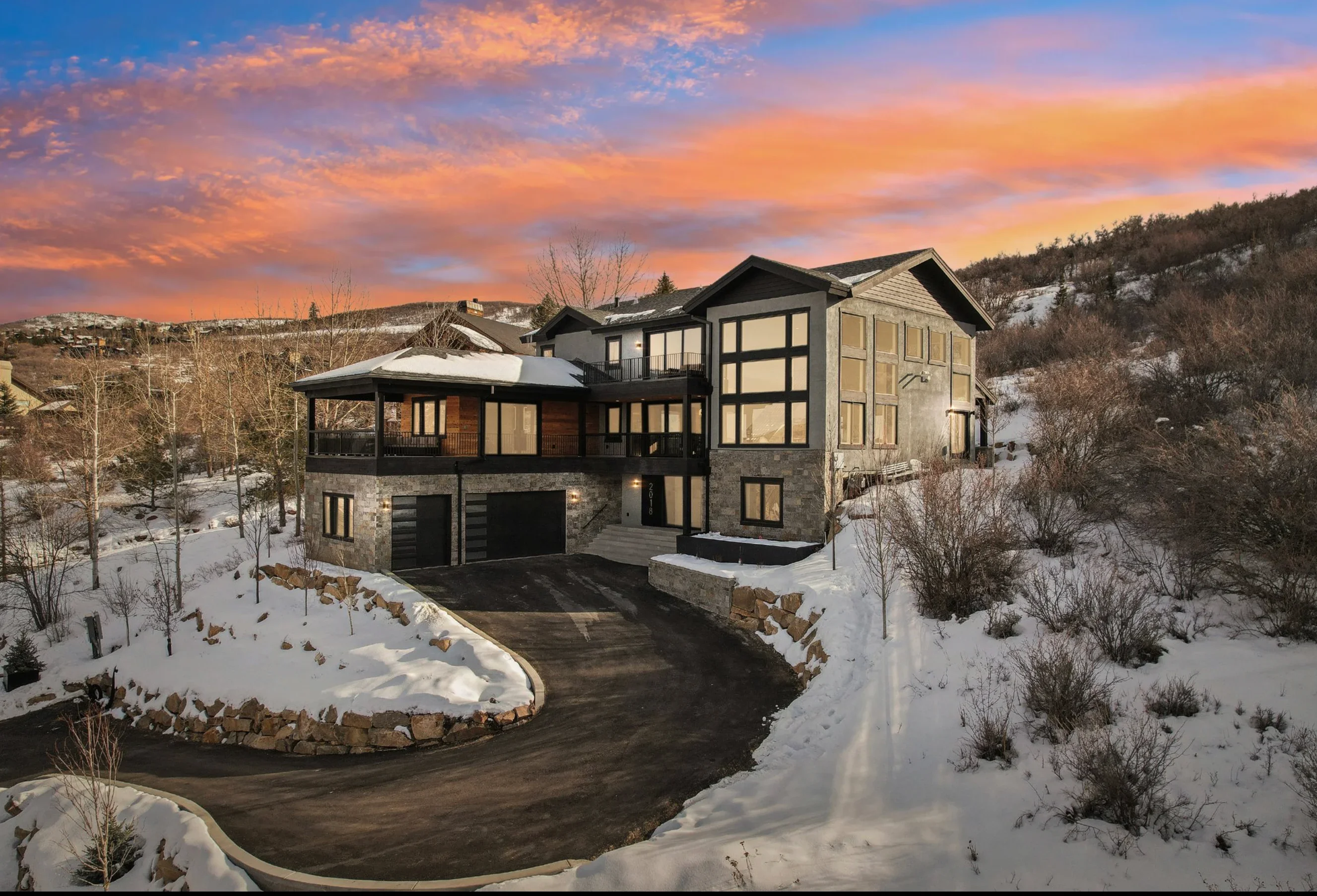 Modern home with stone veneer, dark siding, large windows, and three-car garage on snowy hillside